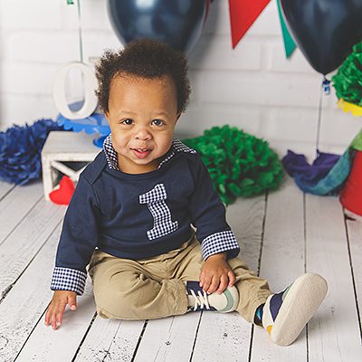a one year old Black baby boy in a navy 1 shirt and khaki pants sits in a colorful photography set - plano cake smash photographer