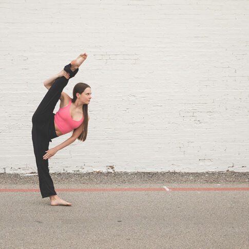 light skinned teenage girl in a pink top and black pants in an elevated leg dance pose in front of a white brick wall - dallas dance photography