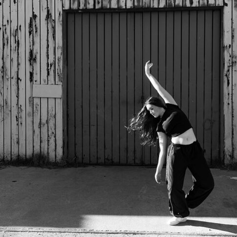 black and white horizontal image of a light skinned teenage girl with long, dark hair in a dance pose, her face slightly down, arm up and hair flying out. She is in front of a warehouse type background - Dallas dance photography