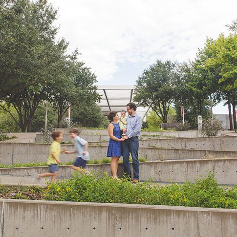 unique family photo in downtown Dallas arts district. Mom, Dad and baby are in the middle of a ramp, while two school age boys are running blurry by them
