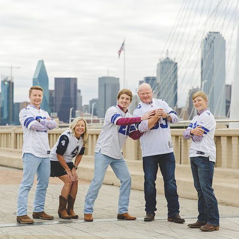 photo of family of five, with mom, dad and three teenage boys, on a bridge with the downtown dallas skyline behind them. They are all wearing dallas cowboys jerseys and looking at the camera - Dallas Family Photography