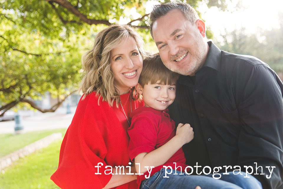 close up image of mother, school age son and father, cuddled up and smiling at the camera, with sunshine and trees behind them. Plano family photography