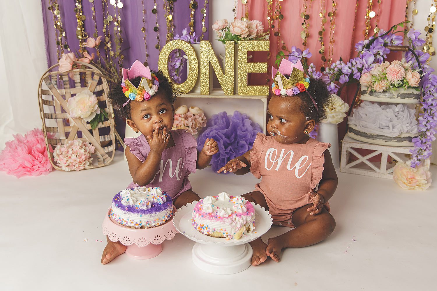 studio set with Black twin one year old girls in pink and purple with cakes, one is putting her hand to her mouth, while the other is looking shocked