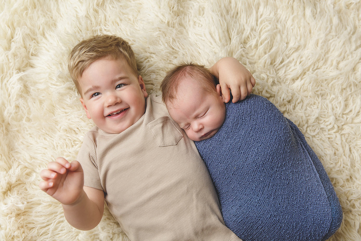 two light skinned boys lying on a cream rug, toddler brother and newborn wrapped in blue