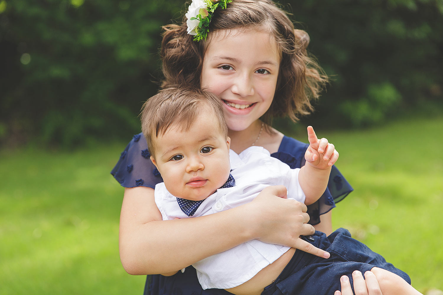 tween girl with light skin and dark hair, holding one year old baby brother with his hand up