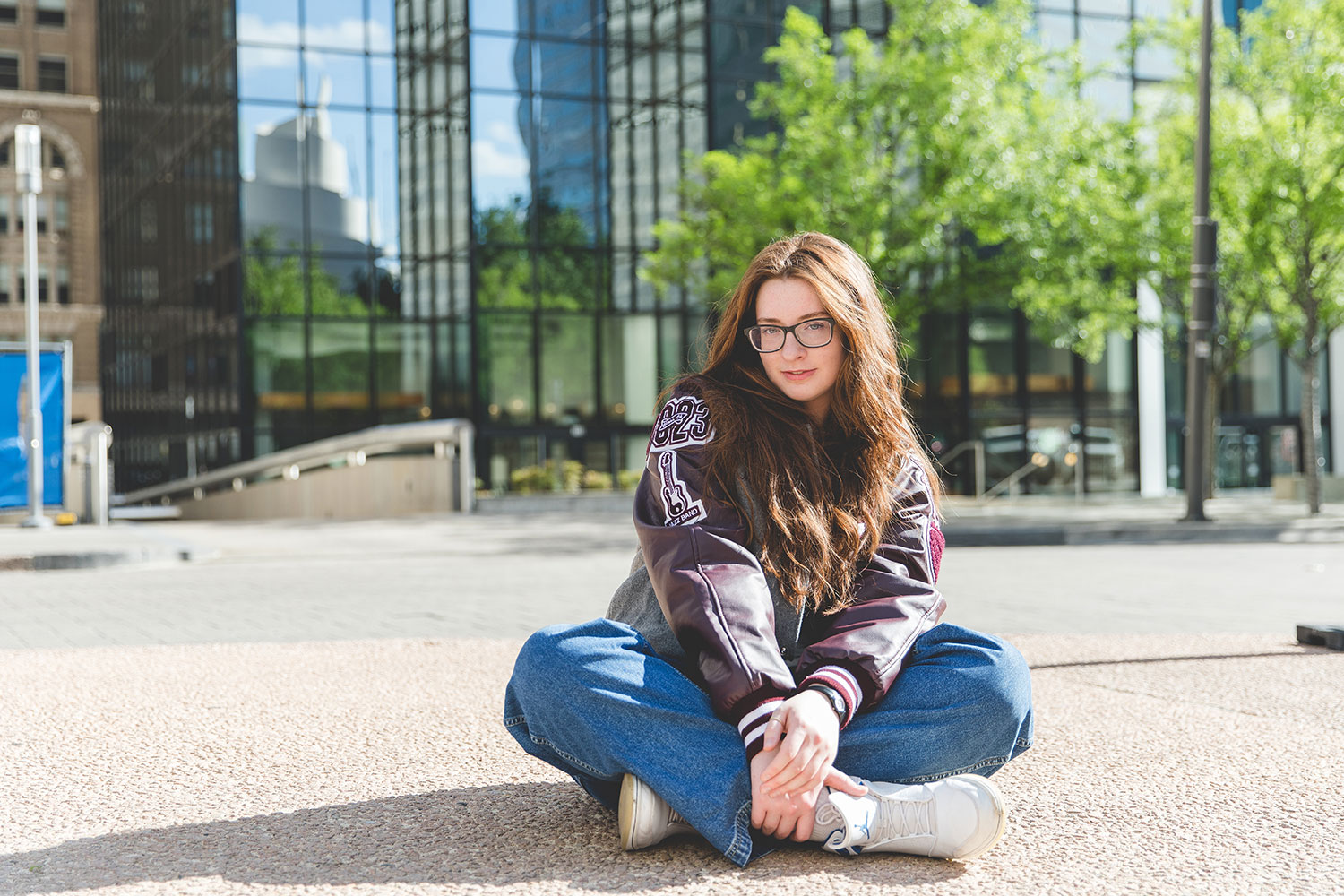 senior girl with long red hair, sitting on the ground in a lettermans jacket and jeans in downtown dallas, with glass buildings behind her - Plano Senior Photographer