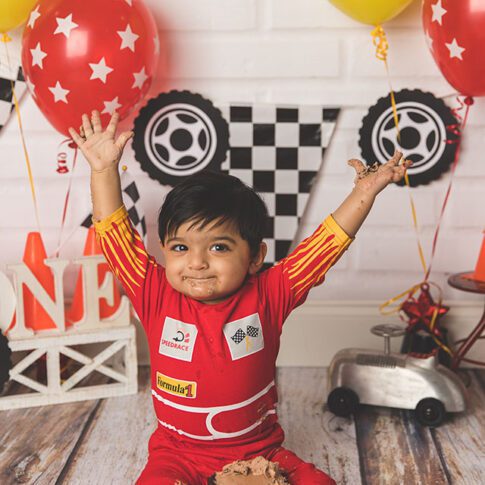 race car themed first birthday cake smash in studio with white brick wall, red yellow, black and white themed decorations and a south asian one year old boy with medium brown skin and black hair, holding his hands in the air in excitement - plano tx photographer