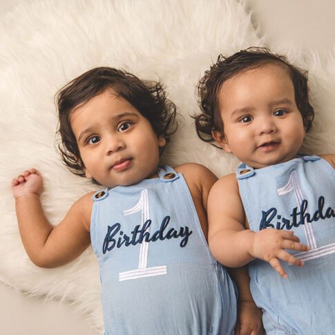 twin one year old boys lying on their back and looking up, on a cream rug wearing blue romper with '1st birthday' on them, they have medium brown skin and dark hair - plano first birthday photographer