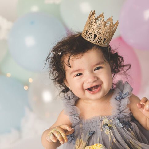 close up image of one year old girl in front of pastel balloon backdrop, she has light skin and curly dark hair, wearing a lopsided gold crown and poofy purple dress - plano cake smash