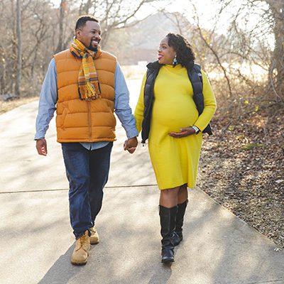 Black couple with a pregnant woman, wearing yellow, blues and orange, looking at one another while walking
