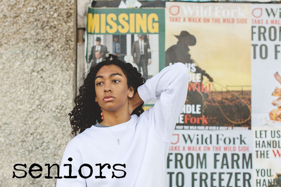 Black senior boy with long, curly hair, in a white sweatshirt with an urban sign background behind him in downtown dallas, senior photography