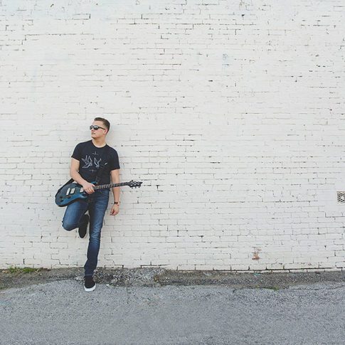 image of senior boy in black shirt and blue jeans, holding a guitar and looking to the left while wearing sunglasses. He is standing against a white brick wall, in Mckinney, TX. Plano Senior Photography
