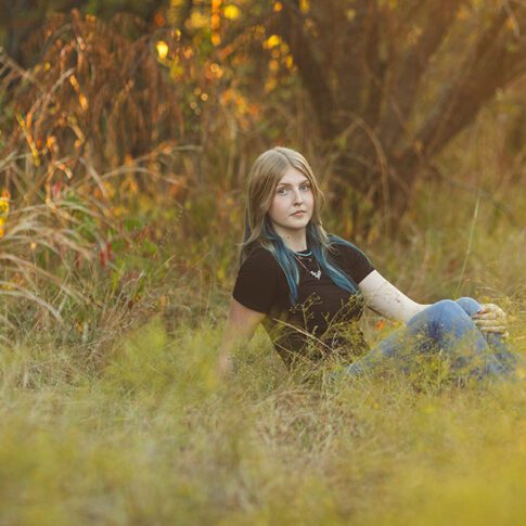photo of a senior girl with light skin sitting in tall grass with warm natural tones behind her - plano senior photography