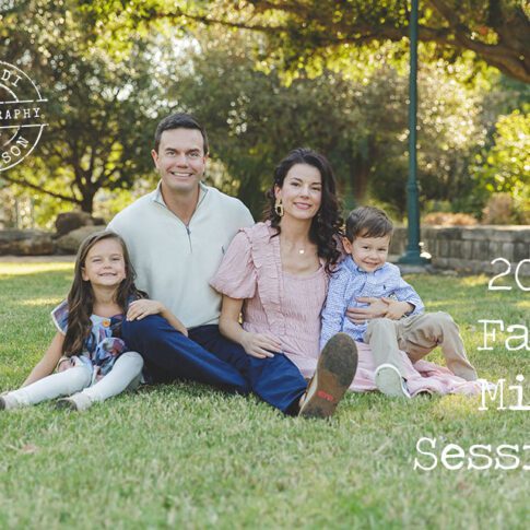 A family of four sitting on the grass with trees behind them, with light skin and brown hair, young school age daughter, dad, mom and preschool age son. Text reads 2023 Fall Mini Sessions.