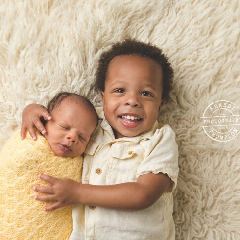 Black toddler boy, and newborn baby, both in yellow, lying on a cream colored rug with big brother smiling at camera